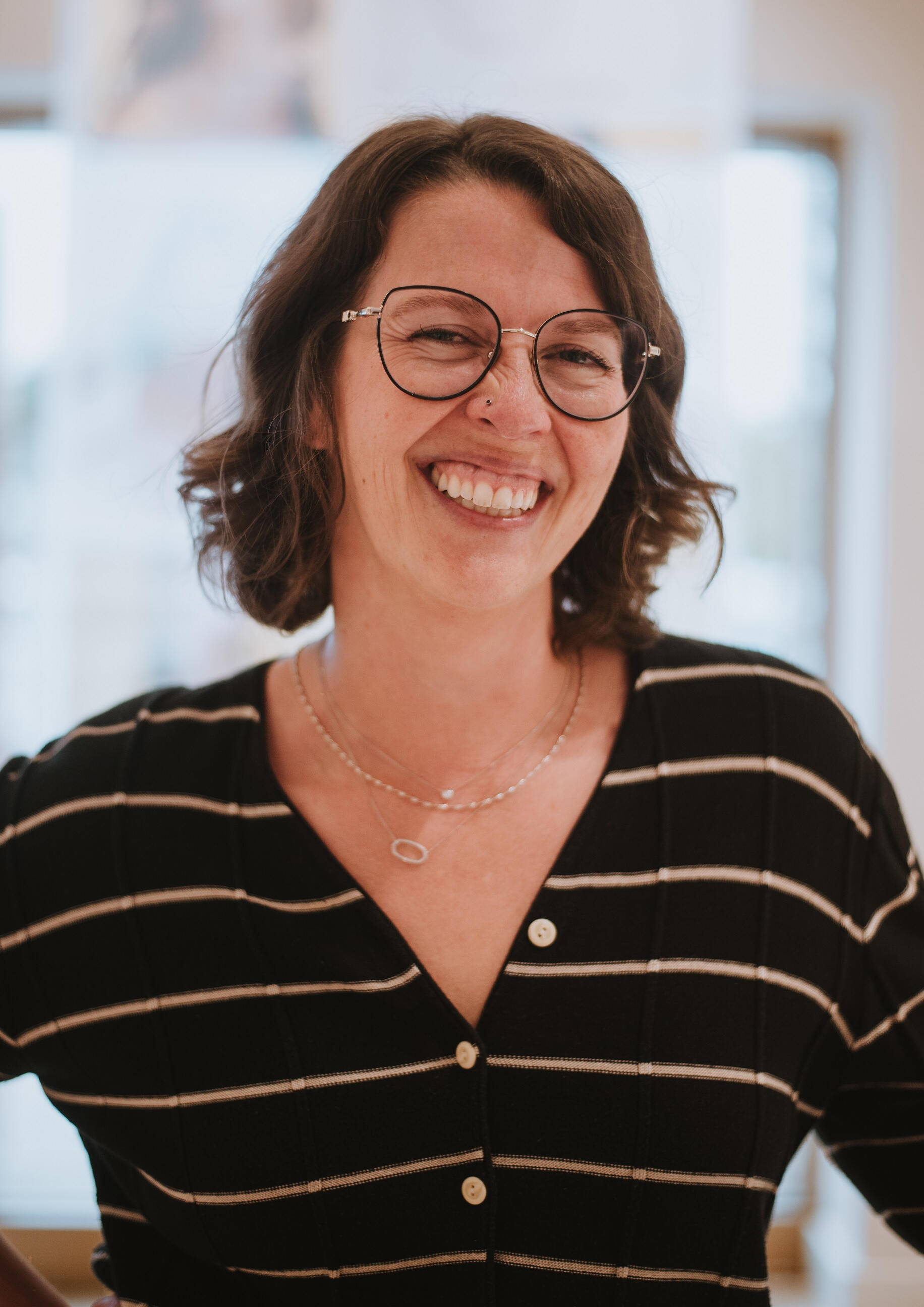 Liz Kardamis LPC, LPAT, 200hr- RYT Headshot of middle aged woman with round, black, thin framed glasses, wavy, chin length brown hair. Her eyes are crinkled on the sides from her large smile. She is wearing a black V-neck sweater with cream stripes and a stack of three thin necklaces.