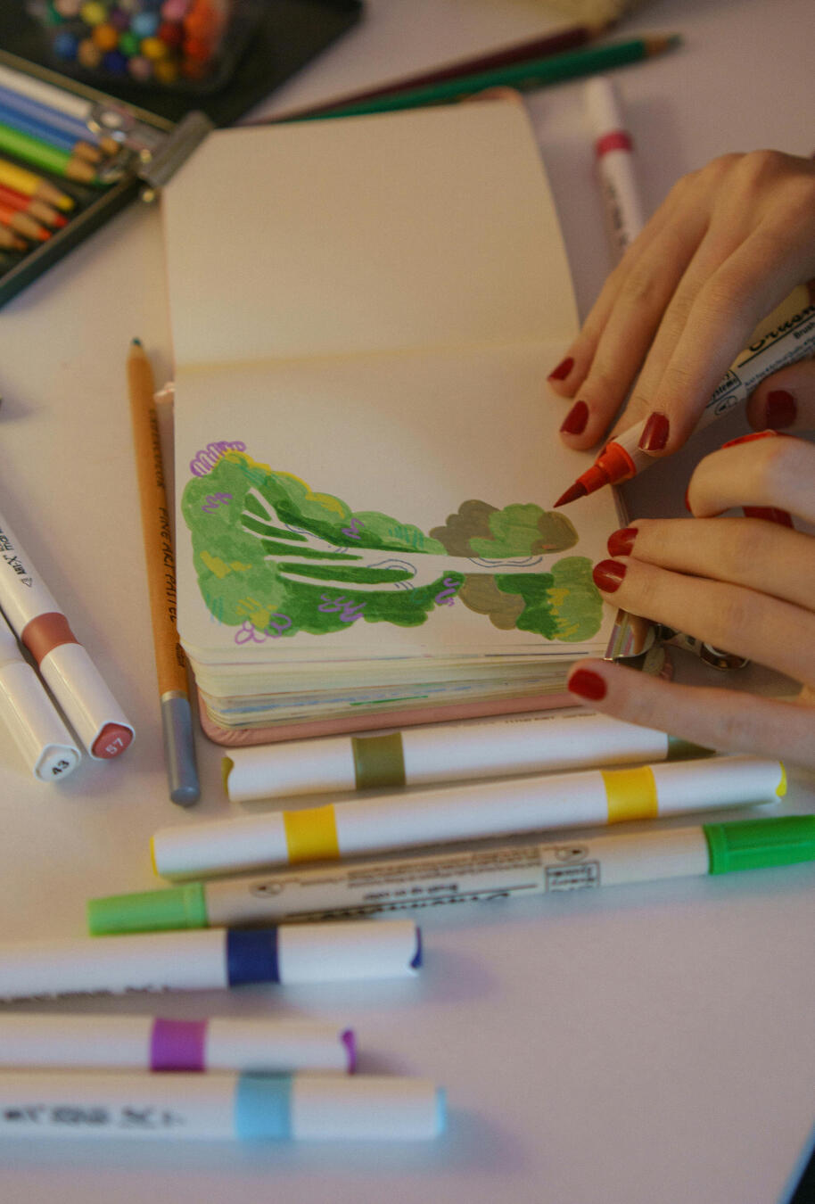 Drawing of tree A close up of an art journal. A woman's fingertips with red painted nails are on the edge of the paper. She is holding a fine tipped red marker over a drawing of a tree with green leaves and grass and blue organic lines on the trunk and branches. Other mar